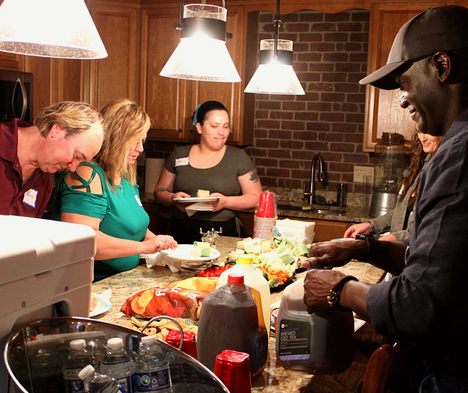 Five people prepare and serve food together in a warmly lit kitchen, surrounded by drinks and snacks.