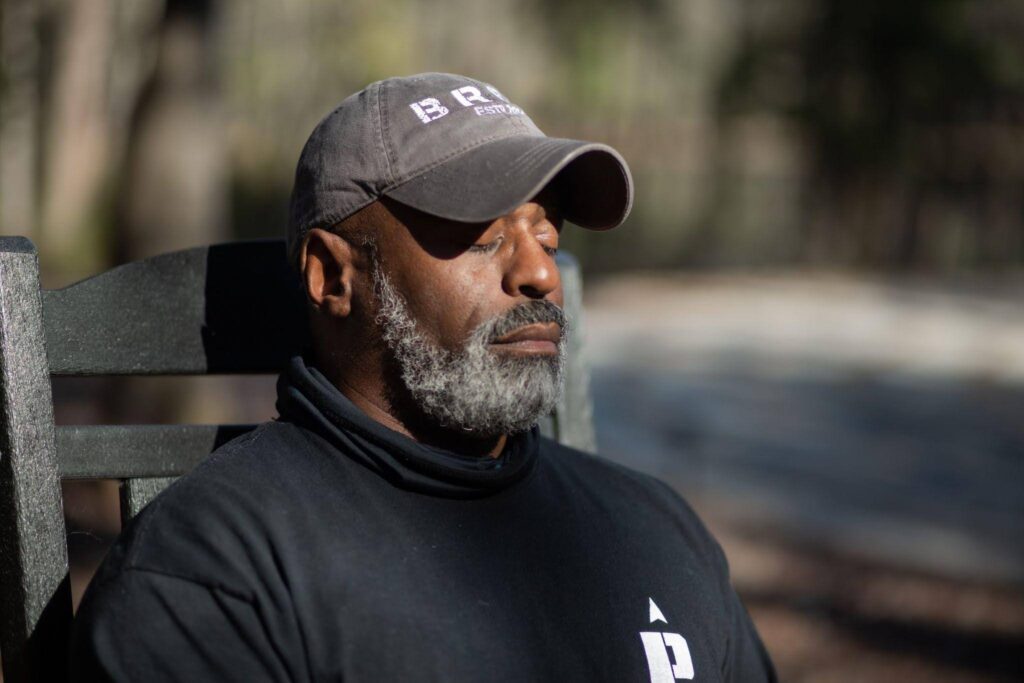 A man with a gray beard and hat sits outdoors with his eyes closed, appearing relaxed in sunlight.
