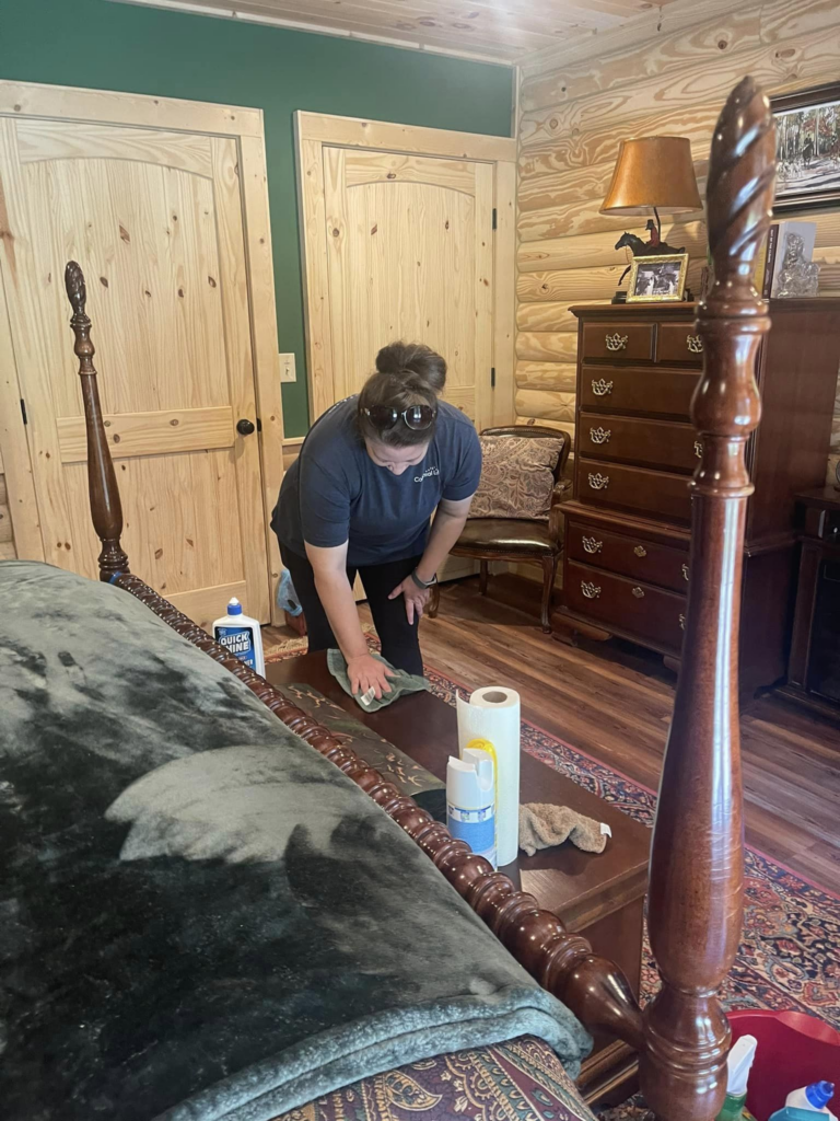 Person cleaning a wooden table in a cozy bedroom with cleaning supplies nearby.