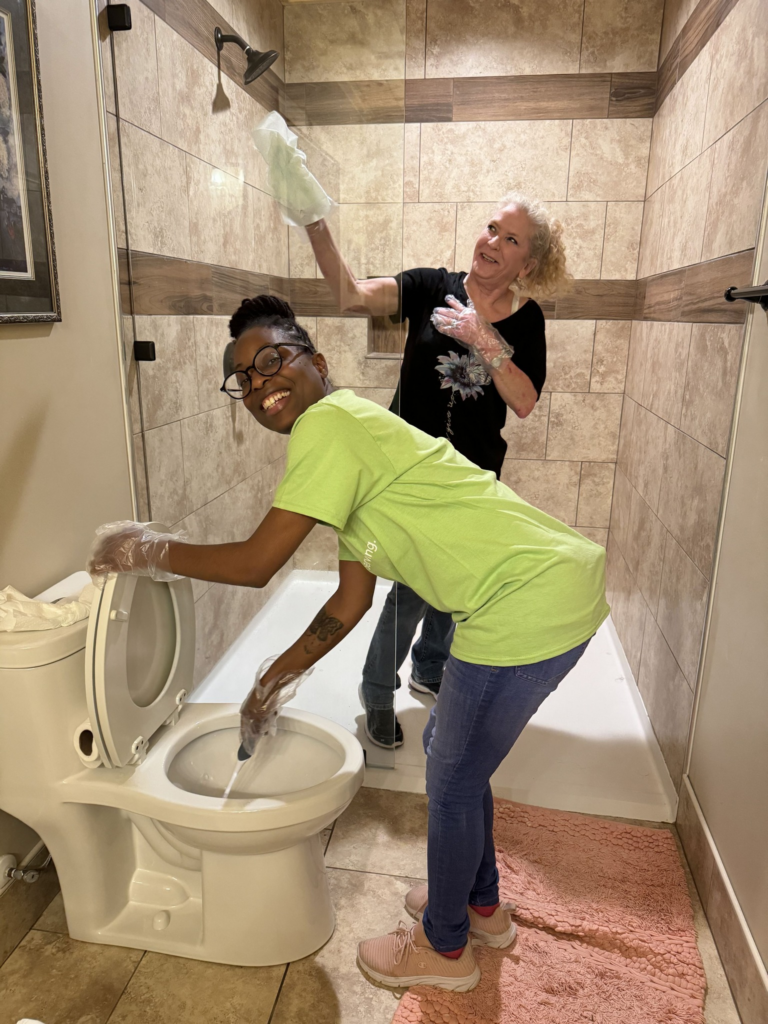 Two women smiling while cleaning a bathroom; one scrubbing a toilet, the other wiping a shower wall.