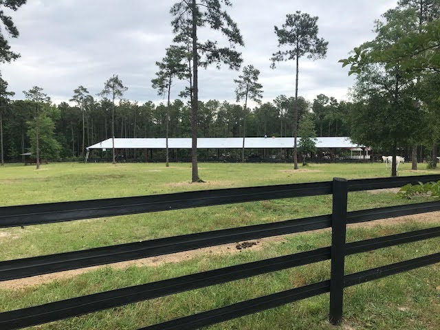 A large barn sits behind a black wooden fence in a grassy, tree-lined rural area under a cloudy sky.