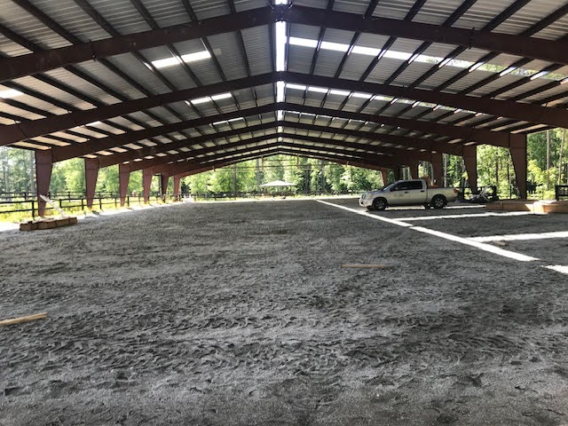 Large open steel structure with a gravel floor, two vehicles parked on the right, and trees visible outside.