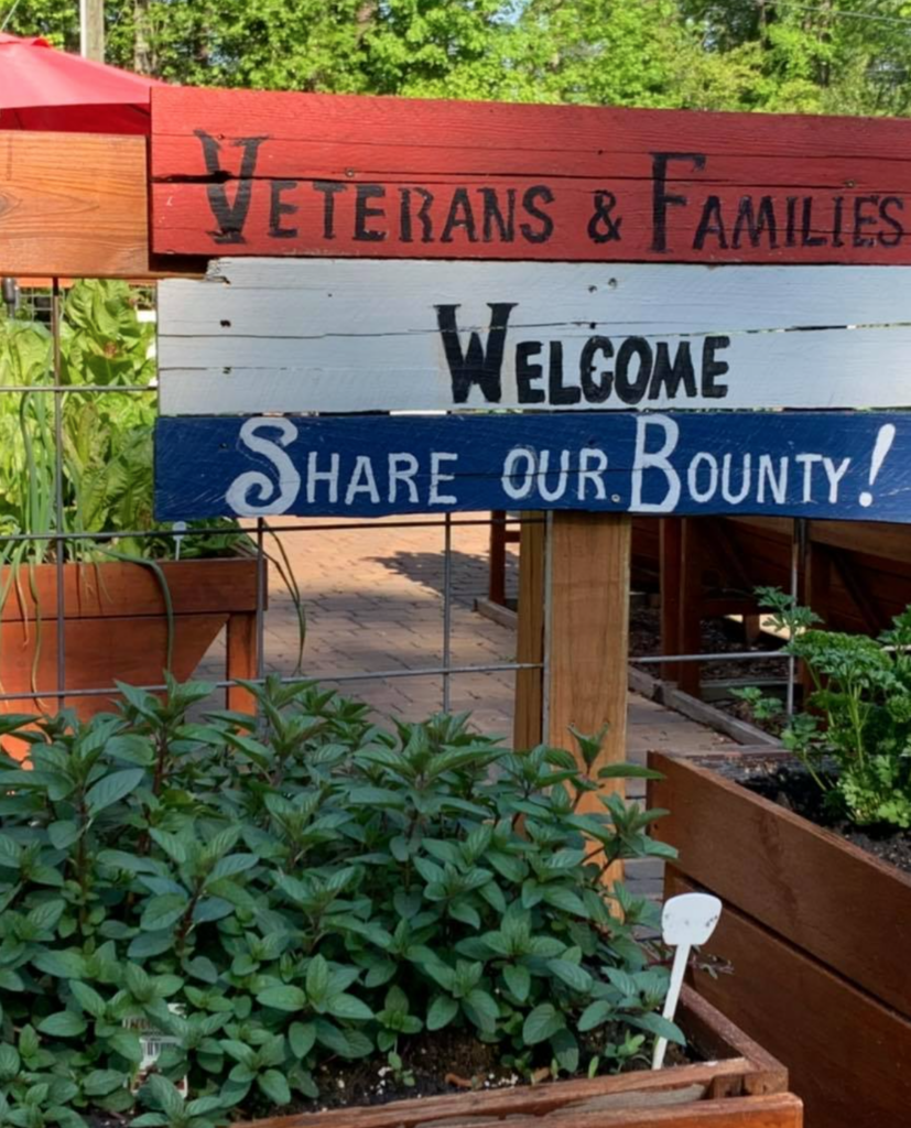 A wooden sign says Veterans & Families Welcome, Share Our Bounty! in a garden with green plants.