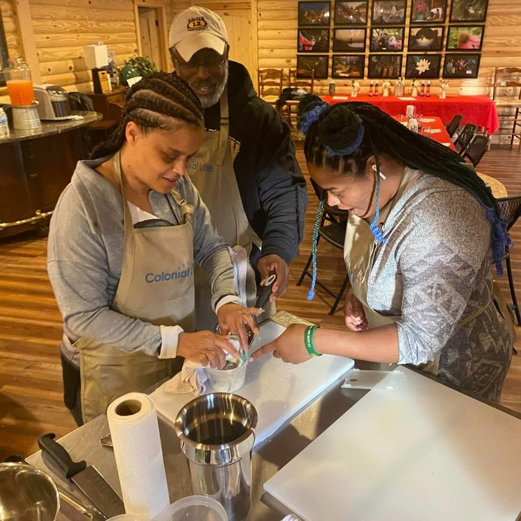 Three people in aprons prepare food together in a cozy kitchen, focusing on a cutting task at the counter.