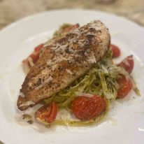 A grilled chicken breast served over pesto pasta with cherry tomatoes and grated cheese on a white plate.