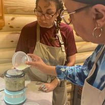 Two women in aprons prepare food, one pours an ingredient into a food processor while the other observes.