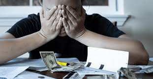 Person with head in hands sits at table covered in bills, papers, and money, appearing stressed.