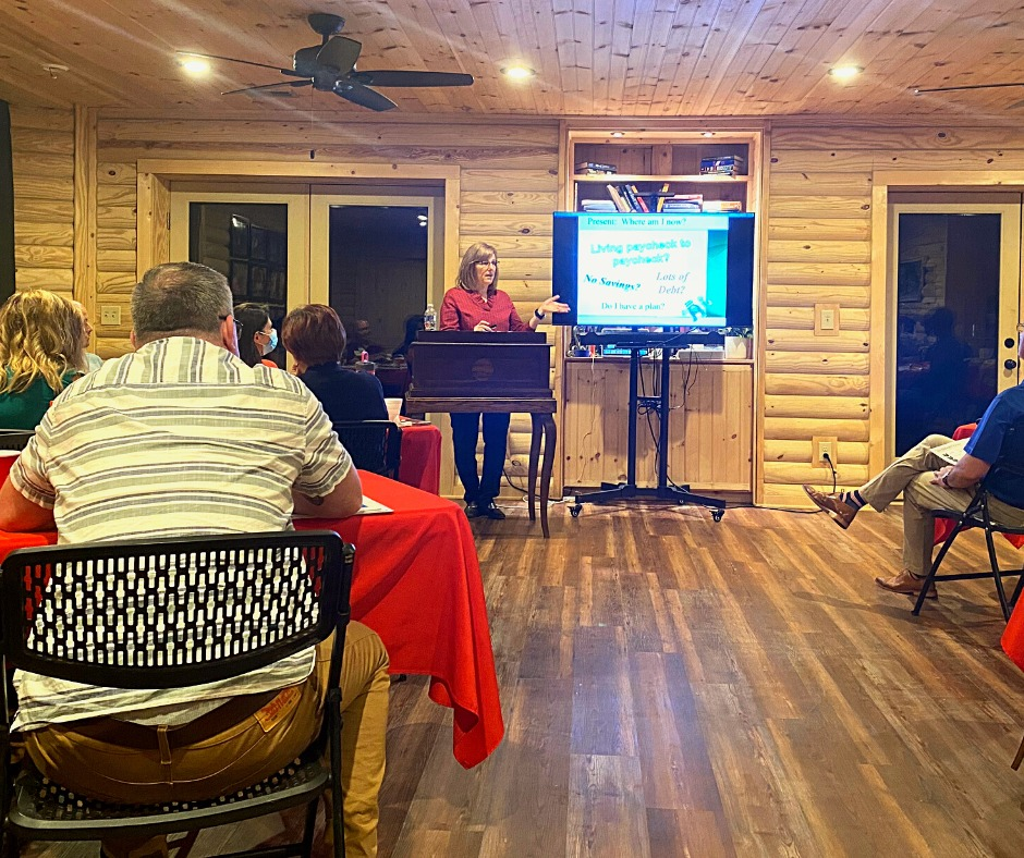 A woman presents a slideshow to a small group seated at tables in a wood-paneled room.