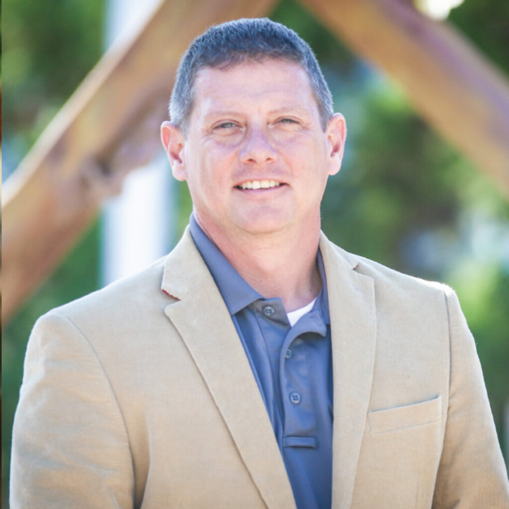 Man in tan blazer and blue shirt smiling outdoors with green blurred background.