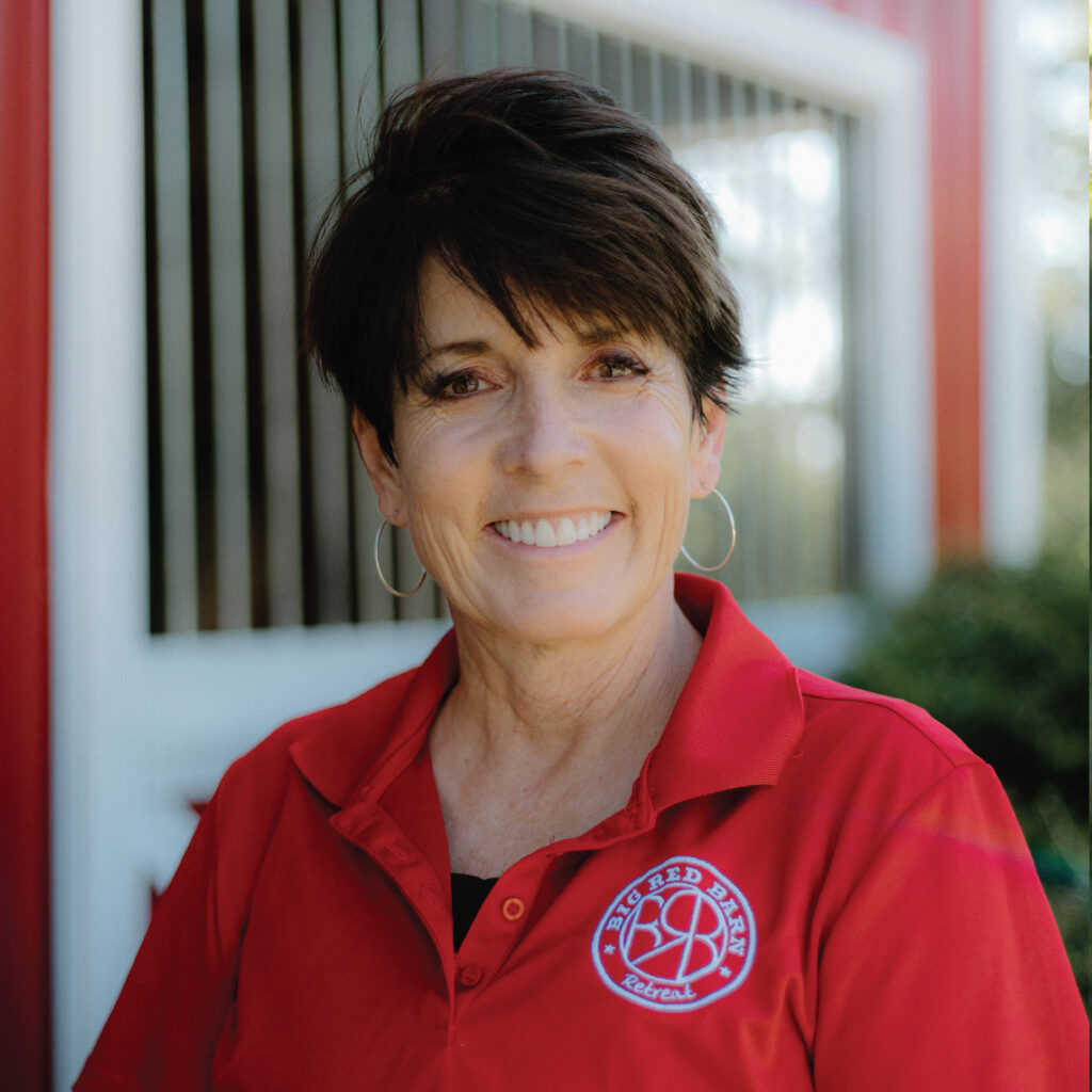 Smiling woman with short dark hair in a red polo shirt standing outdoors in front of a building.