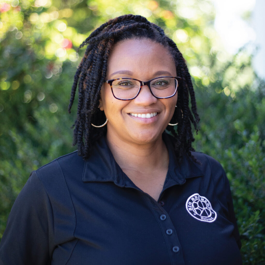 Smiling woman with glasses and braided hair, wearing a black collared shirt, standing outdoors.