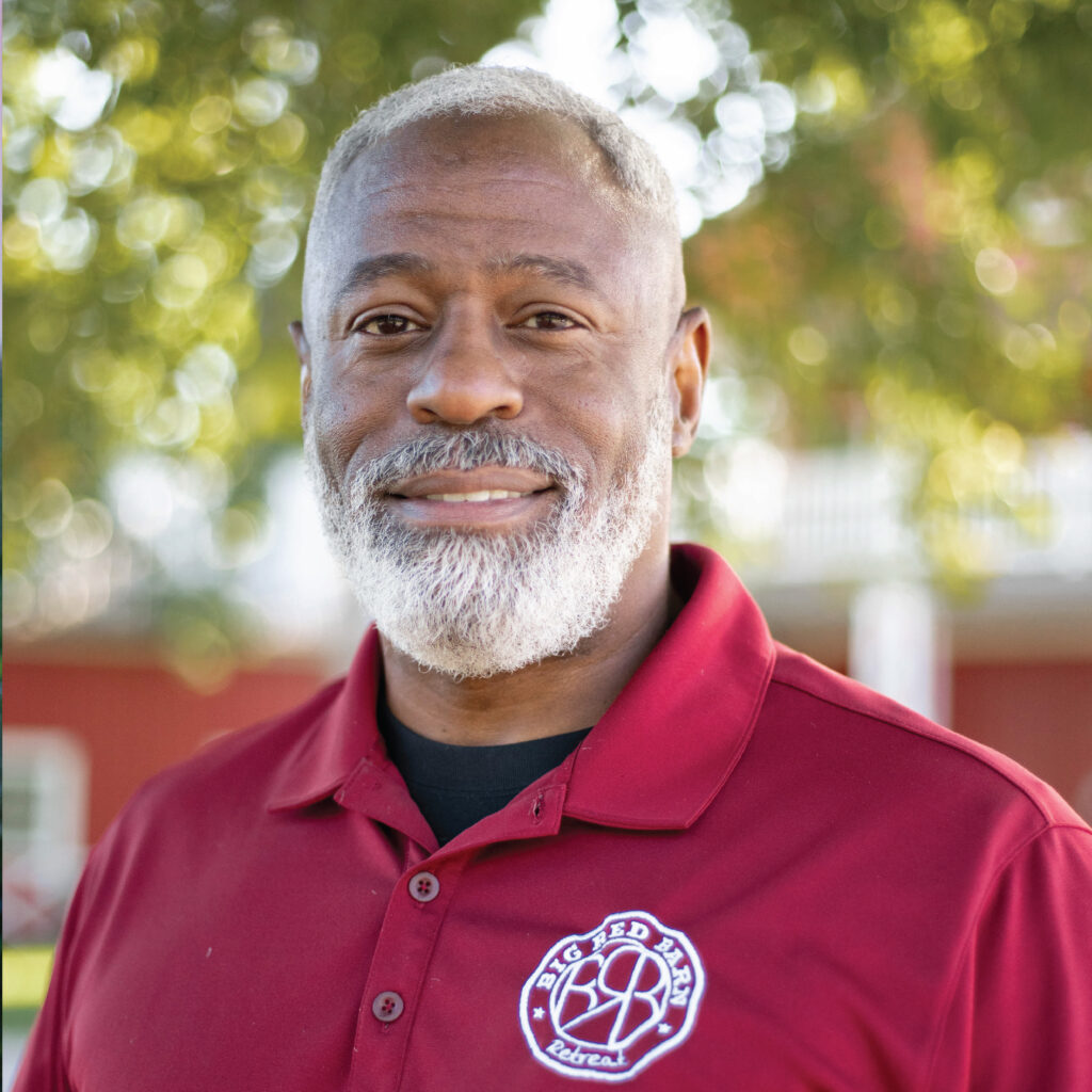 Smiling man with gray beard in red polo shirt stands outdoors in front of trees and a red building.