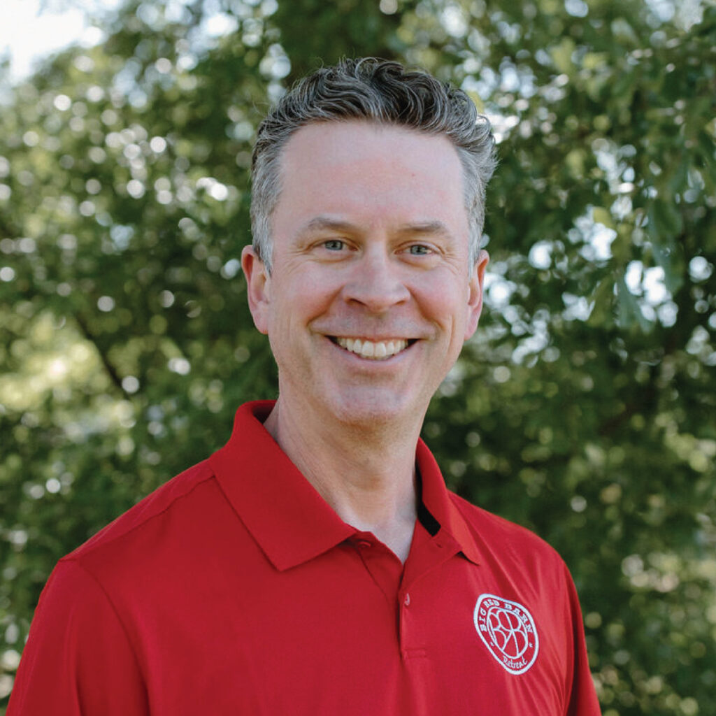 Smiling man in a red polo shirt stands outdoors with green trees in the background.