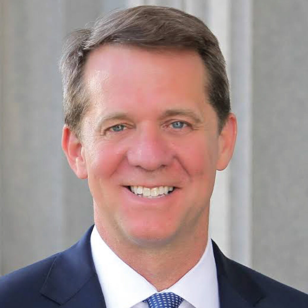 Smiling man in a suit and tie stands in front of a gray, blurred background.