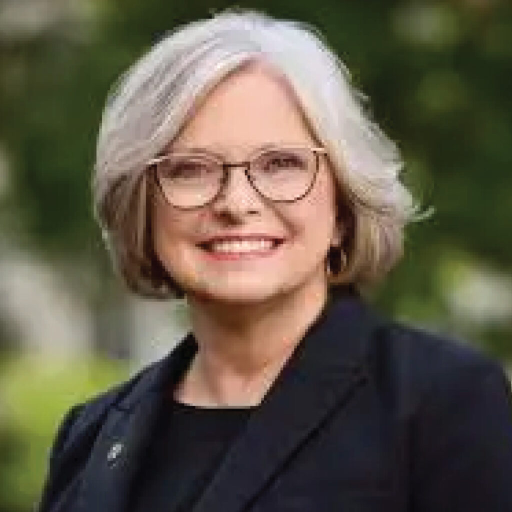 Smiling woman with gray hair, glasses, and a black blazer, outdoors with greenery in the background.