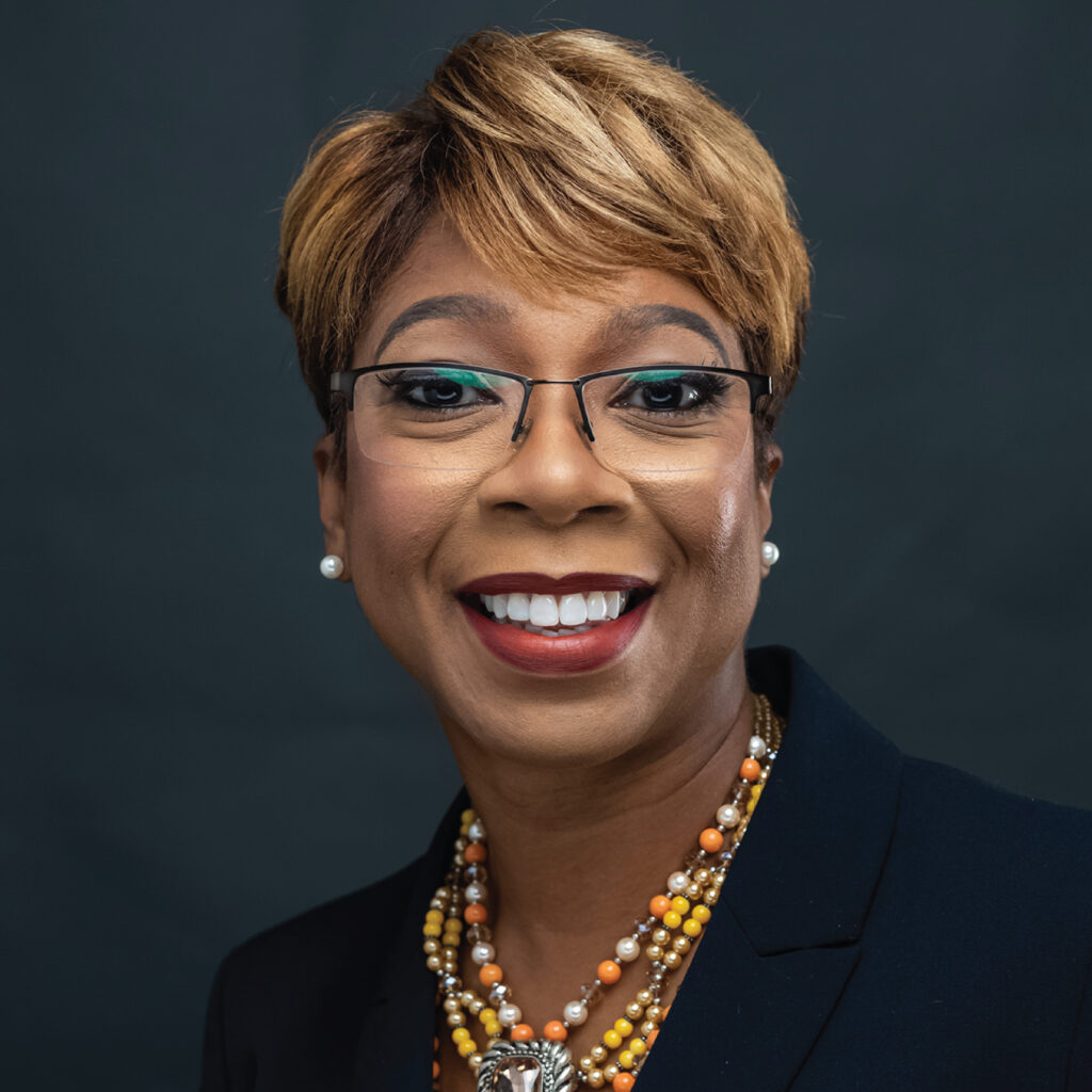 Smiling woman with short hair, glasses, pearl earrings, and a beaded necklace against a dark background.