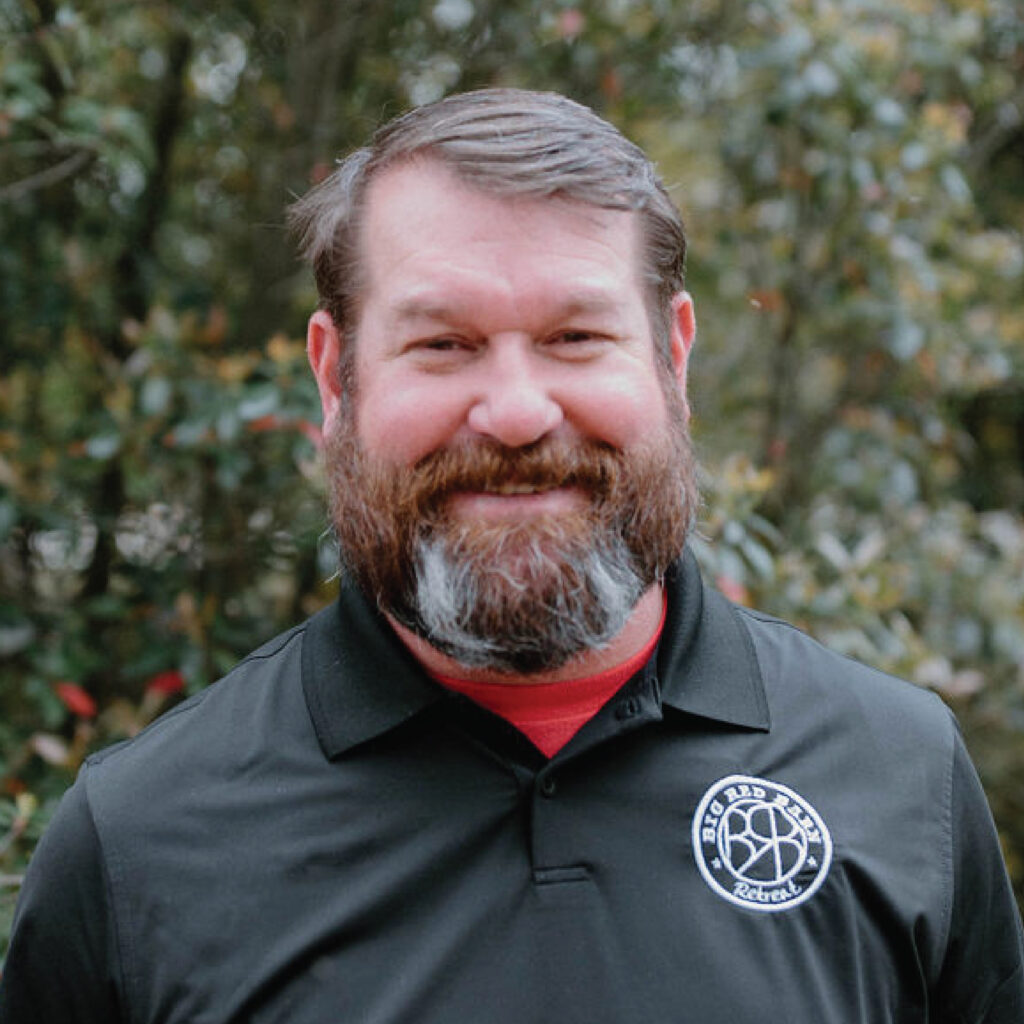 Bearded man smiling outdoors, wearing a black polo shirt with a round white logo on the chest.