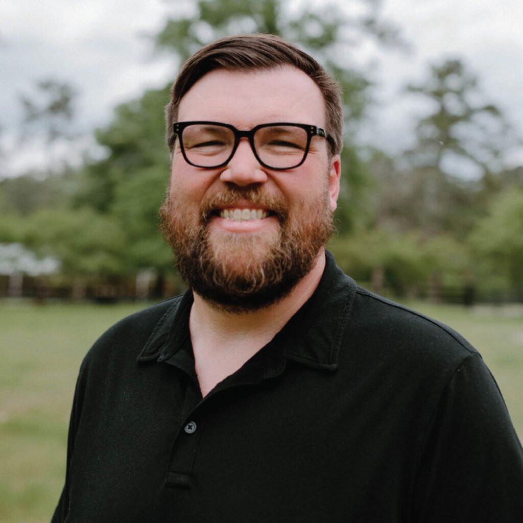 Man with glasses and a beard smiling outdoors, wearing a black collared shirt, with trees in the background.