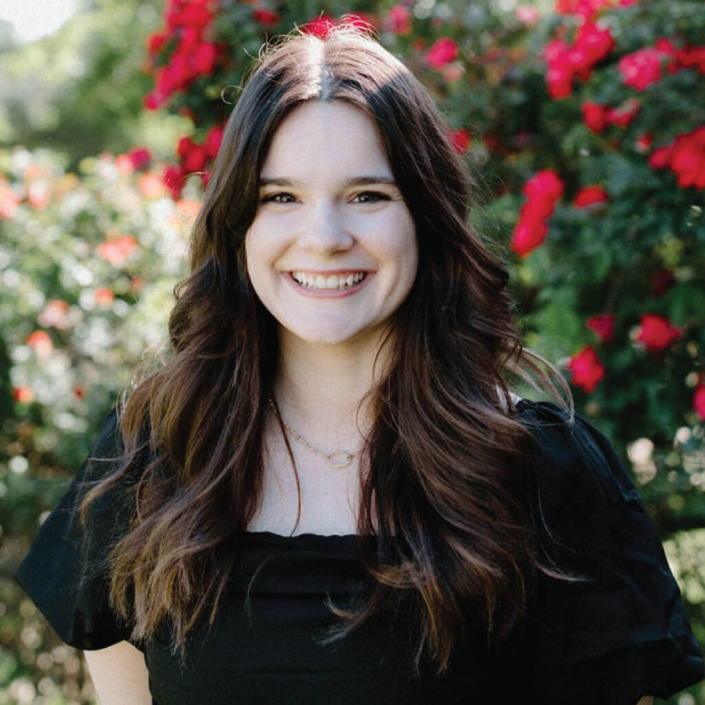 Smiling woman with long brown hair stands in front of vibrant red flowers and greenery, wearing a black top.