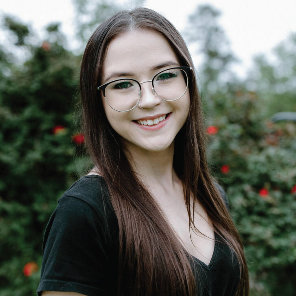Young woman with long brown hair and glasses smiles outdoors, with green foliage in the background.