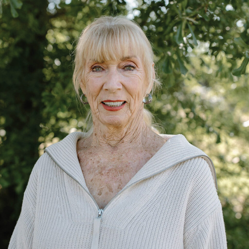 Older woman with light hair and a beige sweater stands outdoors in front of green trees, smiling at the camera.