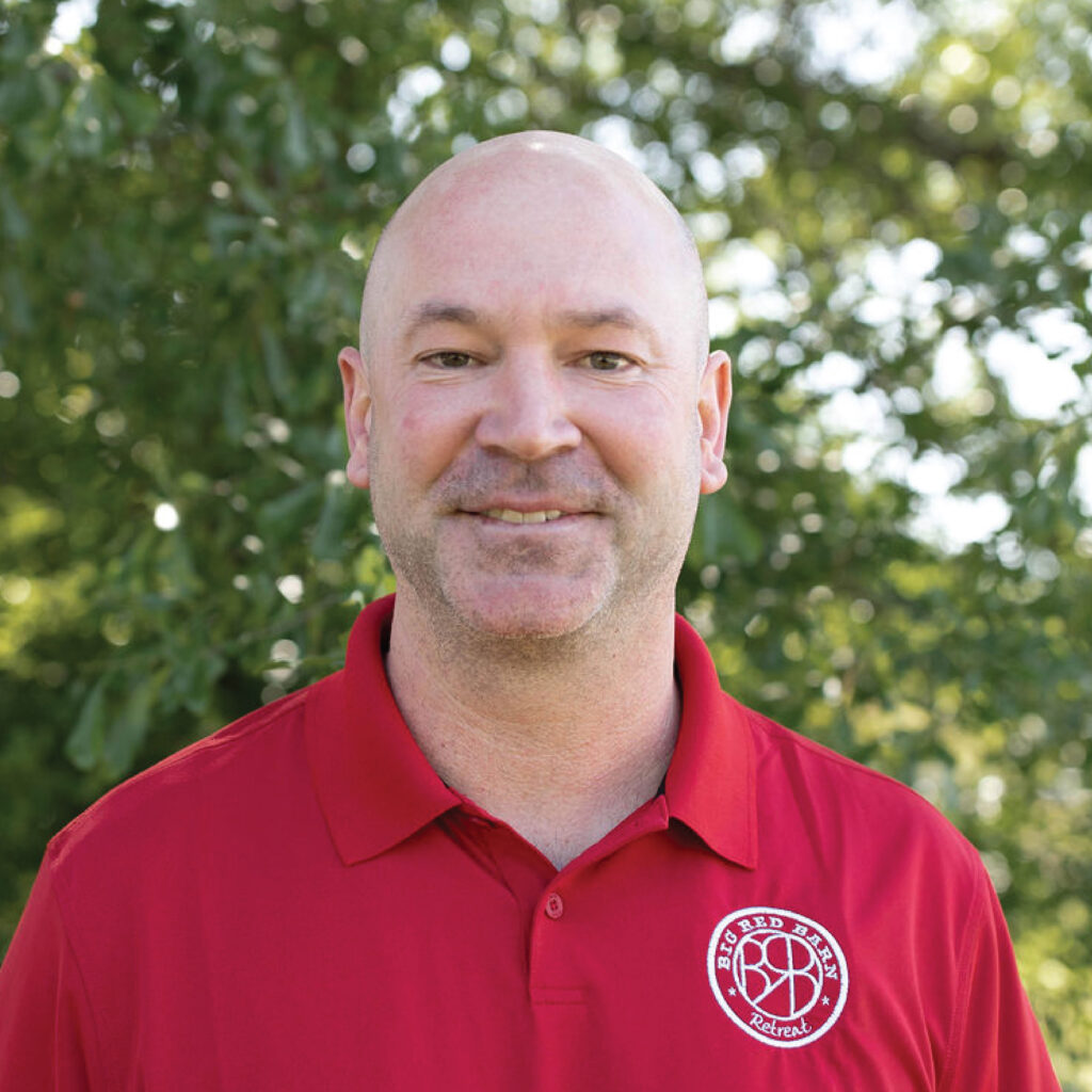A man in a red polo shirt stands outside with trees and sunlight in the background.