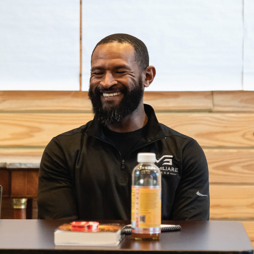 A smiling man with a beard sits at a table with a notebook and a drink in front of him.