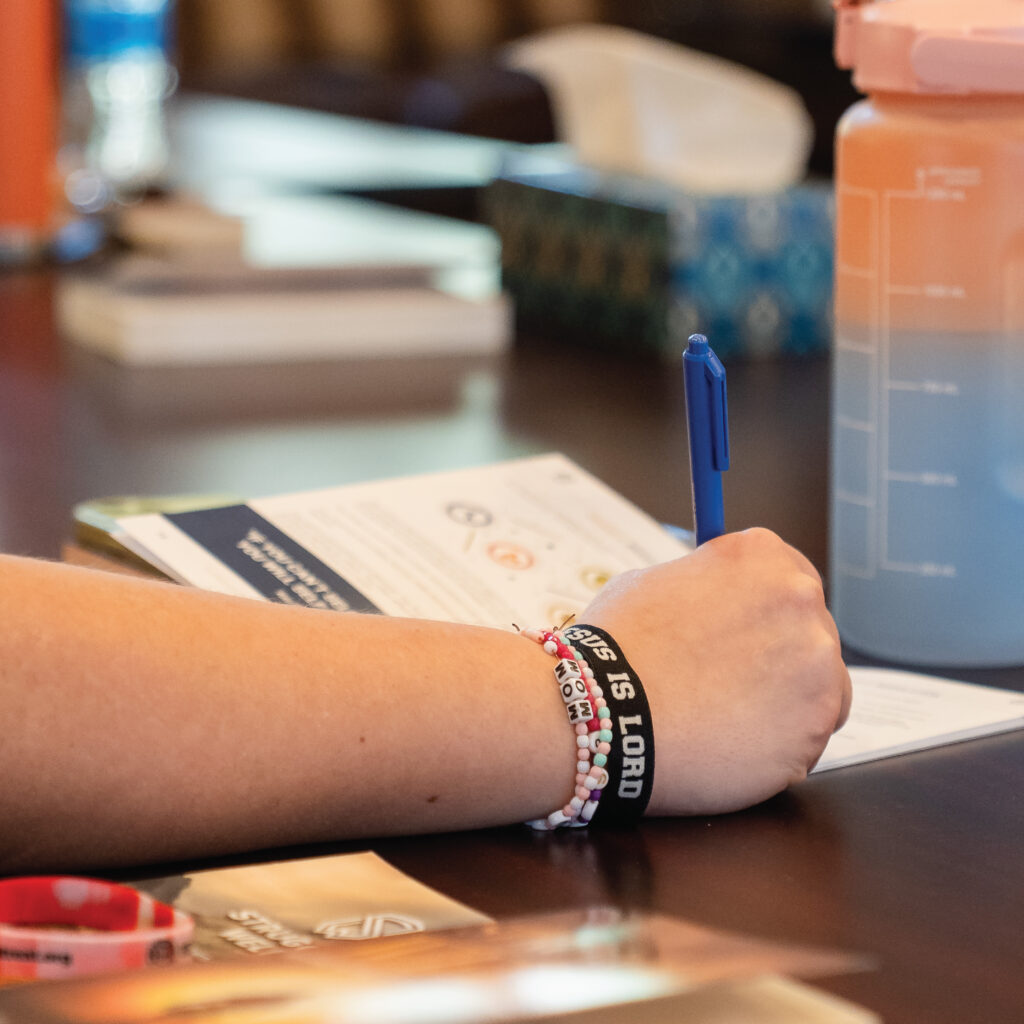 A person wearing a “JESUS IS LORD” bracelet writes in a notebook next to a large water bottle on a table.