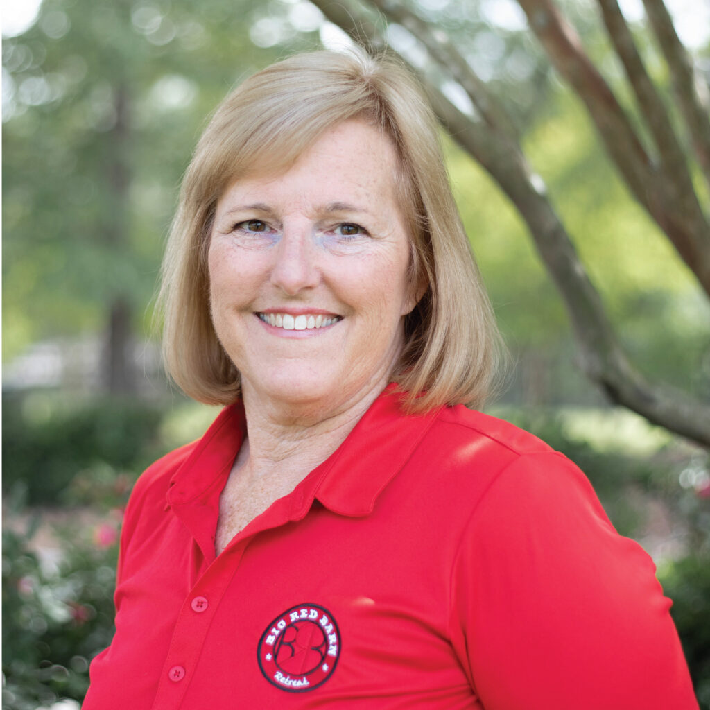 Smiling woman with blonde hair in a red polo shirt standing outdoors, trees blurred in the background.