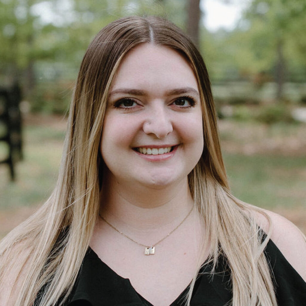 A woman with long, straight, blonde hair smiles outdoors with trees and greenery in the background.