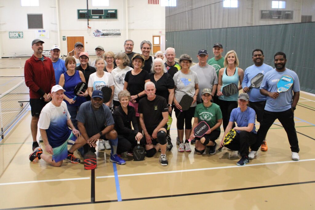 A group of adults pose with pickleball paddles on an indoor court, smiling at the camera.