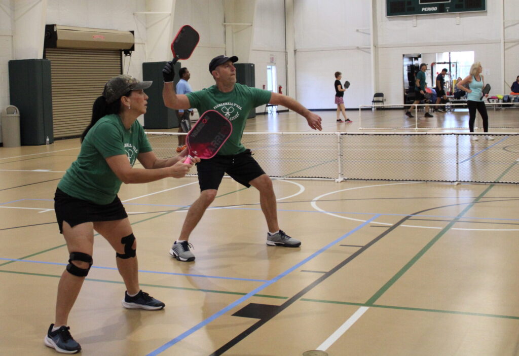 Two people play pickleball indoors, both wearing green shirts and holding paddles, ready to hit the ball.