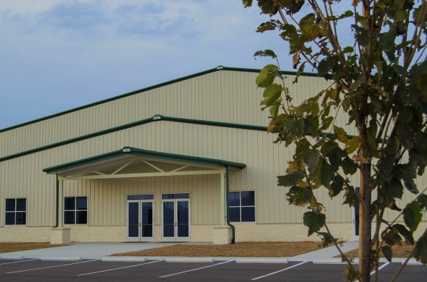 Large beige metal building with green trim, glass double doors, and empty parking spaces; tree in foreground.