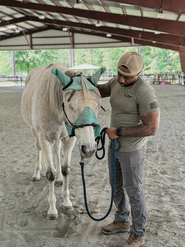 A man gently petting a white horse wearing a fly mask under a covered riding arena.