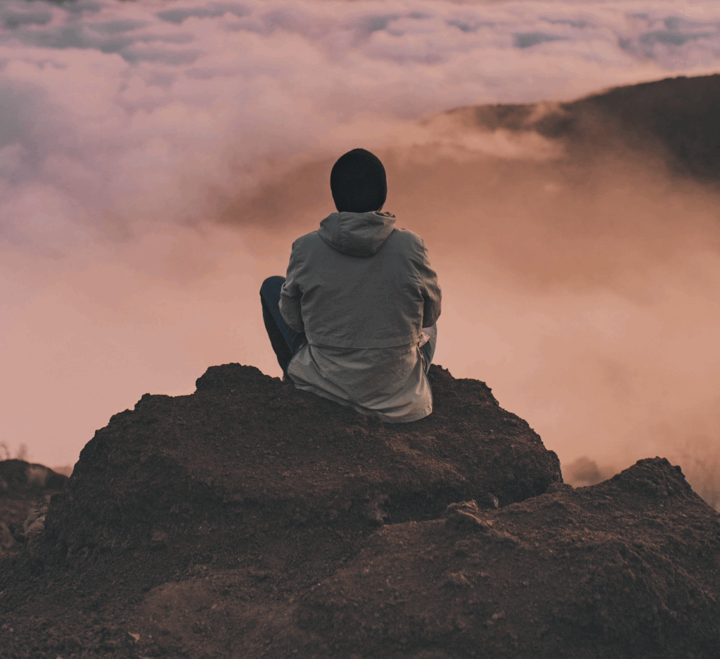 Person sitting on a rocky hilltop, looking at clouds and a colorful sky during sunset or sunrise.