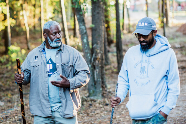 Two men walking in a forest, talking and holding walking sticks on a sunny day.