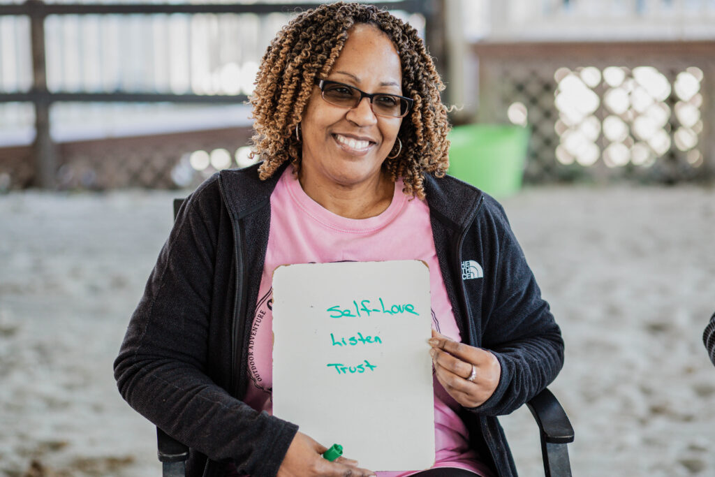 Smiling woman holding a whiteboard with “Self-Love, Listen, Trust” written on it.