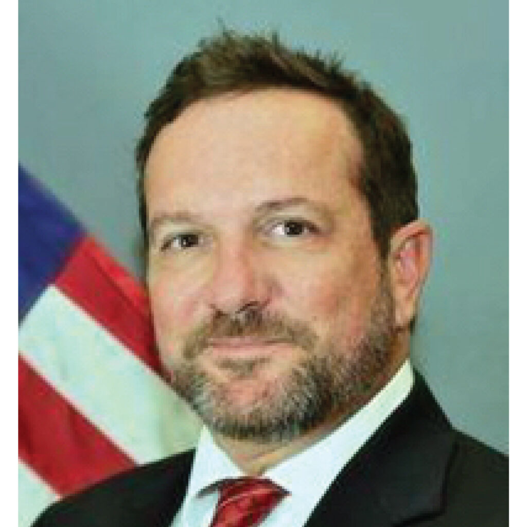 A man in a suit and red tie poses in front of a U.S. flag, looking at the camera.