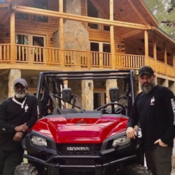 Two men in black shirts pose with a red Honda ATV in front of a large log cabin-style house.