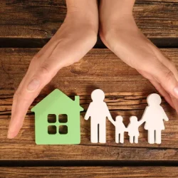 Hands protect paper cutouts of a family and a house on a wooden surface.